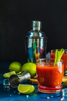 Tomato juice with celery, spices, salt and ice in portion drink glasses with copy space. Bloody Mary cocktail. Alcoholic drink and ingredients at dark blue concrete table surface.