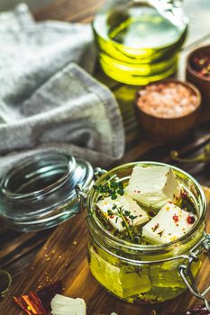 Feta cheese marinated in olive oil with fresh herbs in glass jar. Wooden background.