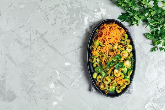 Delicious roasted Brussels sprouts and marinated carrot chips in served on black plate, light gray concrete table surface, top view, copy space for you text.