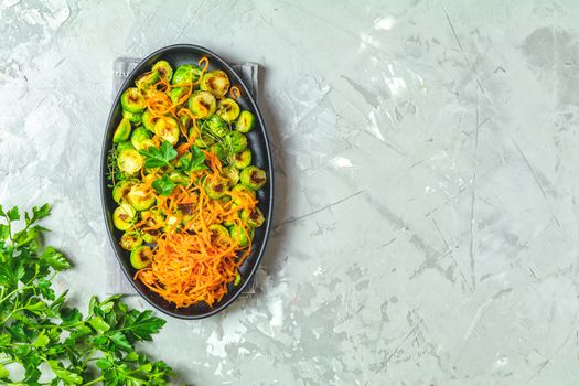 Delicious roasted Brussels sprouts and marinated carrot chips in served on black plate, light gray concrete table surface, top view, copy space for you text.