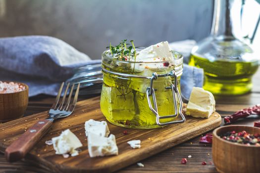 Feta cheese marinated in olive oil with fresh herbs in glass jar. Wooden background.