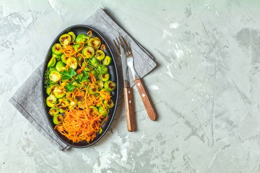 Delicious roasted Brussels sprouts and marinated carrot chips in served on black plate, light gray concrete table surface, top view, copy space for you text.