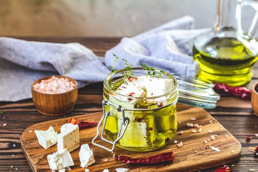 Feta cheese marinated in olive oil with fresh herbs in glass jar. Wooden background.