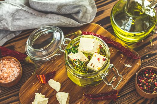 Feta cheese marinated in olive oil with fresh herbs in glass jar. Wooden background.