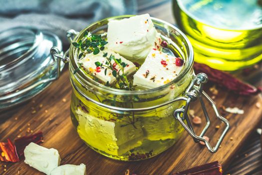 Feta cheese marinated in olive oil with fresh herbs in glass jar. Wooden background