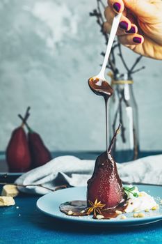 Chocolate sauce pours from a spoon on red pears in wine. Red pears are in a blue plate. The concept of still life on blue concrete surface.