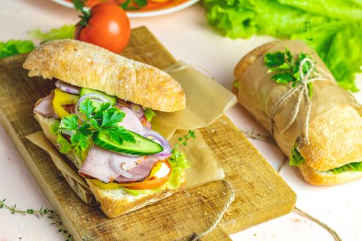 Italian porchetta sandwich. Ciabatta sandwich with ham, tomato, cheese, pepper, onion and salad on wooden cutting board with ingredients.