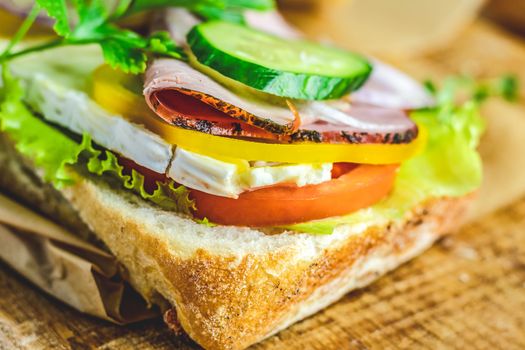 Ciabatta sandwich with ham, tomato, cheese, pepper, onion and salad on wooden cutting board with ingredients.