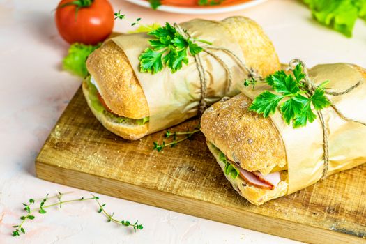 Ciabatta sandwich with ham, tomato, cheese, pepper, onion and salad on wooden cutting board with ingredients.