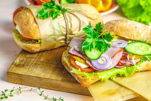 Ciabatta sandwich with ham, tomato, cheese, pepper, onion and salad on wooden cutting board with ingredients.