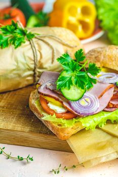 Ciabatta sandwich with ham, tomato, cheese, pepper, onion and salad on wooden cutting board with ingredients.