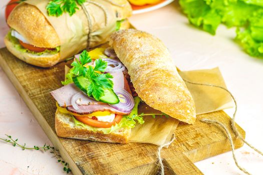 Ciabatta sandwich with ham, tomato, cheese, pepper, onion and salad on wooden cutting board with ingredients.