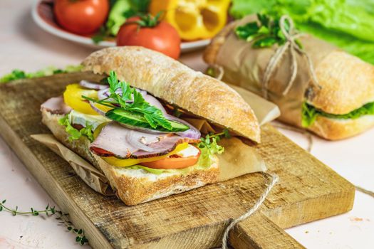 Ciabatta sandwich with ham, tomato, cheese, pepper, onion and salad on wooden cutting board with ingredients.