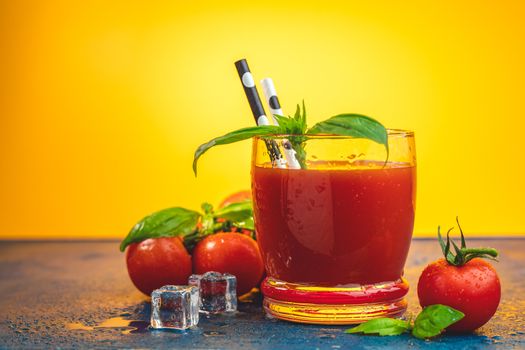 Red cocktail with tomato juice between tomatoes, fresh basil and ice. Delicious tomato bloody mary cocktail on dark blue concrete table with spot light. Yellow background.