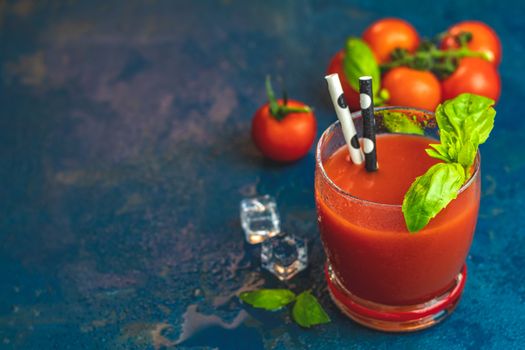 Red cocktail with tomato juice between tomatoes, fresh basil and ice. Delicious tomato bloody mary cocktail on dark blue concrete table.