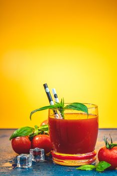 Red cocktail with tomato juice between tomatoes, fresh basil and ice. Delicious tomato bloody mary cocktail on dark blue concrete table with spot light. Yellow background.