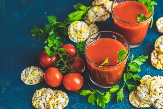 Red cocktail with tomato juice between tomatoes, basil, parsley and nutritious cereal breads. Delicious tomato bloody mary cocktail on dark blue concrete table.