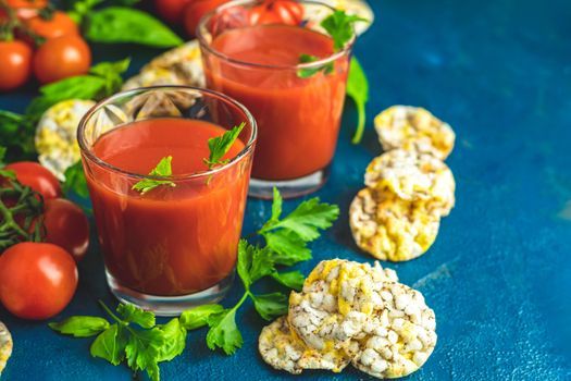 Red cocktail with tomato juice between tomatoes, basil, parsley and nutritious cereal breads. Delicious tomato bloody mary cocktail on dark blue concrete table.