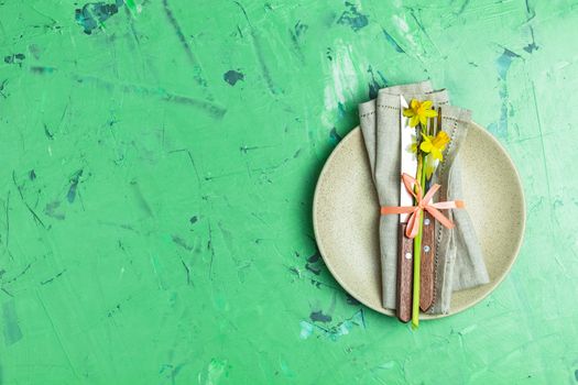 Empty beige plate and cutlery with daffodils on a napkin. Top view, green concrete surface background, copy space for you text