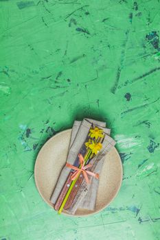 Empty beige plate and cutlery with daffodils on a napkin. Top view, green concrete surface background, copy space for you text