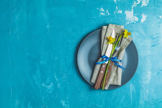 Empty blue plate and cutlery with daffodils on a napkin. Top view, green concrete surface background, copy space for you text
