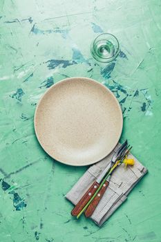 Empty beige plate and cutlery with daffodils on a napkin. Top view, green concrete surface background, copy space for you text