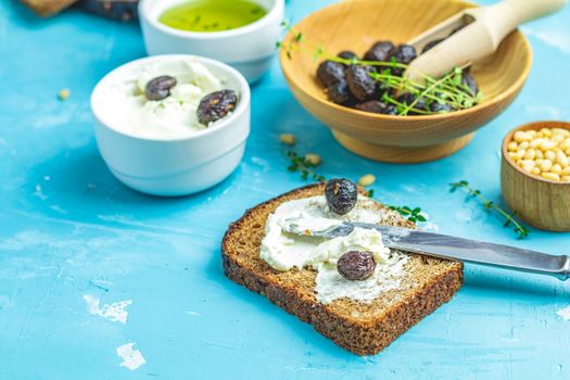 Cream Cheese on a slice of bread topped, bowl of cream cheese, mozzarella, pine nuts and traditional greek italian appetizer dried olives on blue concrete table surface.