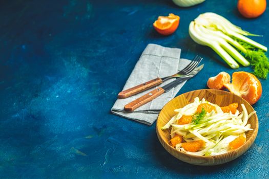 Traditional sicilian italian salad with fennel and tangerine. Fennel and orange citrus salad on wooden plate between ingredients on dark blue concrete table surface. Modern, light, raw, veg cuisine