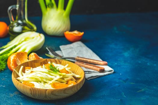 Traditional sicilian italian salad with fennel and tangerine. Fennel and orange citrus salad on wooden plate between ingredients on dark blue concrete table surface. Modern, light, raw, veg cuisine