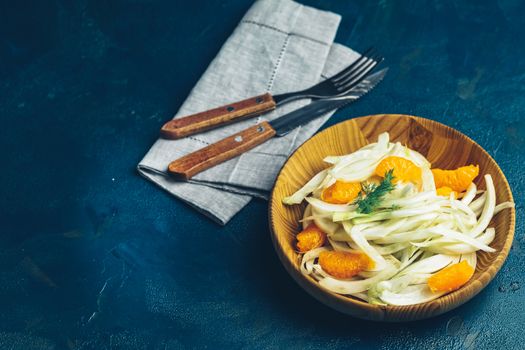 Traditional sicilian italian salad with fennel and tangerine. Fennel and orange citrus salad on wooden plate between ingredients on dark blue concrete table surface. Modern, light, raw, veg cuisine