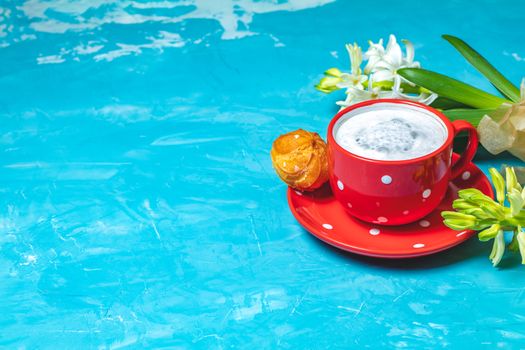 Red in white dotted cup of coffee with milk, delicious profiteroles with cream and white hyacinths on blue concrete surface background, copy space. Beautiful spring greeting card.