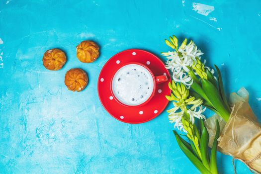 Red in white dotted cup of coffee with milk, delicious profiteroles with cream and white hyacinths on blue concrete surface background. Top view, copy space. Beautiful spring greeting card.