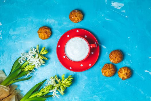 Red in white dotted cup of coffee with milk, delicious profiteroles with cream and white hyacinths on blue concrete surface background. Top view, copy space. Beautiful spring greeting card.