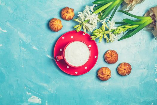 Red in white dotted cup of coffee with milk, delicious profiteroles with cream and white hyacinths on blue concrete surface background. Top view, copy space. Beautiful spring greeting card.