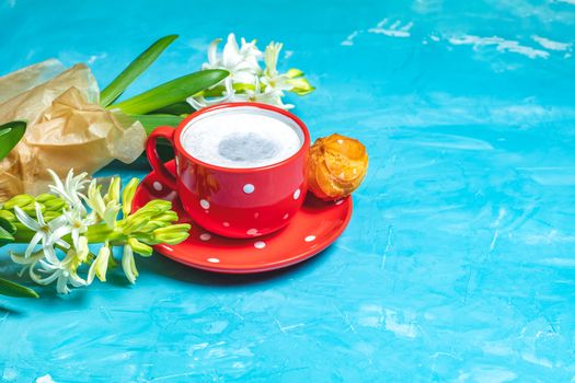 Red in white dotted cup of coffee with milk, delicious profiteroles with cream and white hyacinths on blue concrete surface background, copy space. Beautiful spring greeting card.