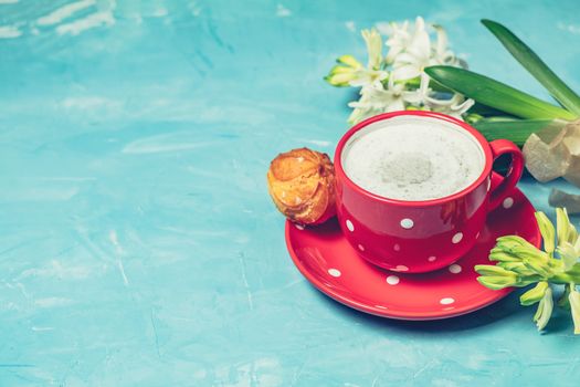 Red in white dotted cup of coffee with milk, delicious profiteroles with cream and white hyacinths on blue concrete surface background, copy space. Beautiful spring greeting card.