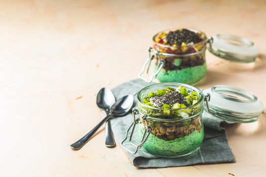 Two chia seed pudding with matcha green tea, kiwi and granola, orange in glasses on light pink concrete background. Healthy breakfast. Square image