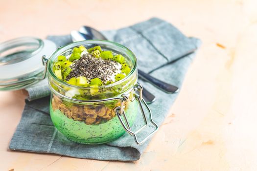 Chia seed pudding with matcha green tea, kiwi and granola in glass on light pink peachy concrete background. Healthy breakfast.