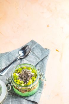 Chia seed pudding with matcha green tea, kiwi and granola in glass on light pink peachy concrete background. Healthy breakfast.
