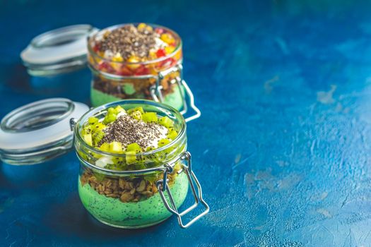 Two chia seed pudding with matcha green tea, kiwi and granola, orange in glasses on dark blue concrete background. Healthy breakfast.