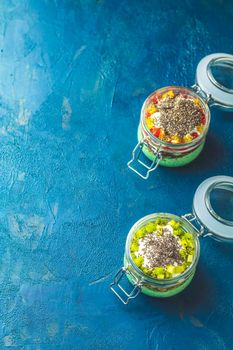 Two chia seed pudding with matcha green tea, kiwi and granola, orange in glasses on dark blue concrete background. Healthy breakfast.