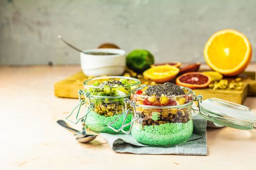 Two chia seed pudding with matcha green tea, kiwi and granola, orange in glasses on light pink concrete background. Healthy breakfast.