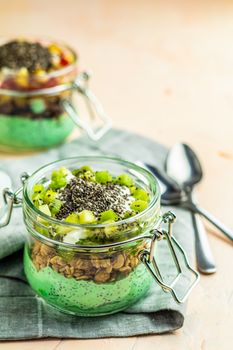 Two chia seed pudding with matcha green tea, kiwi and granola, orange in glasses on light pink concrete background. Healthy breakfast. 