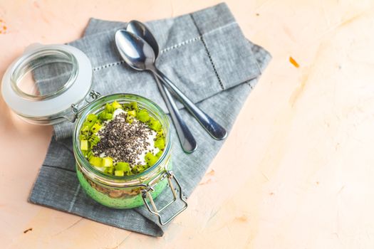 Chia seed pudding with matcha green tea, kiwi and granola in glass on light pink peachy concrete background. Healthy breakfast.