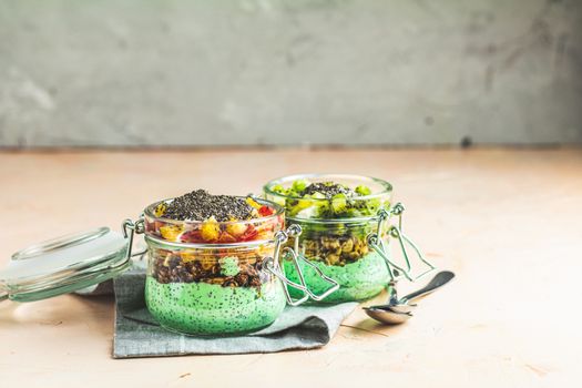 Two chia seed pudding with matcha green tea, kiwi and granola, orange in glasses on light pink concrete background. Healthy breakfast. 