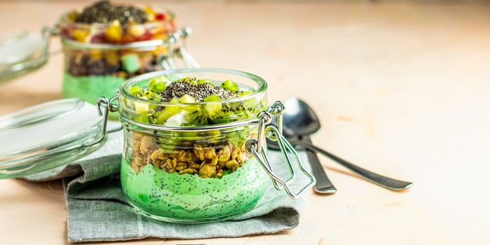 Two chia seed pudding with matcha green tea, kiwi and granola, orange in glasses on light pink concrete background. Healthy breakfast. Image banner format