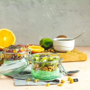 Two chia seed pudding with matcha green tea, kiwi and granola, orange in glasses on light pink concrete background. Healthy breakfast. Square image