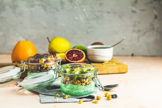 Two chia seed pudding with matcha green tea, kiwi and granola, orange in glasses on light pink concrete background. Healthy breakfast. 