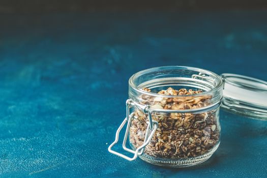 Open glass jar of organic granola with berries, coconut chips and seeds on a dark blue concrete table surface.