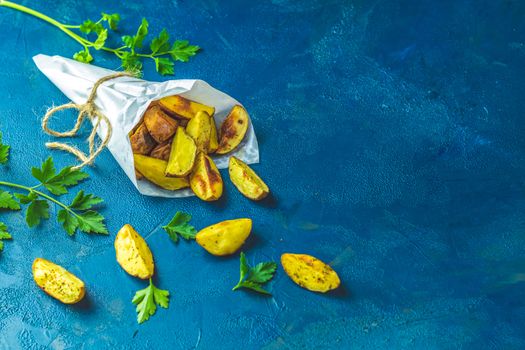 Baked potato wedges on paper with addition sea salt and parsley on a dark blue concrete background